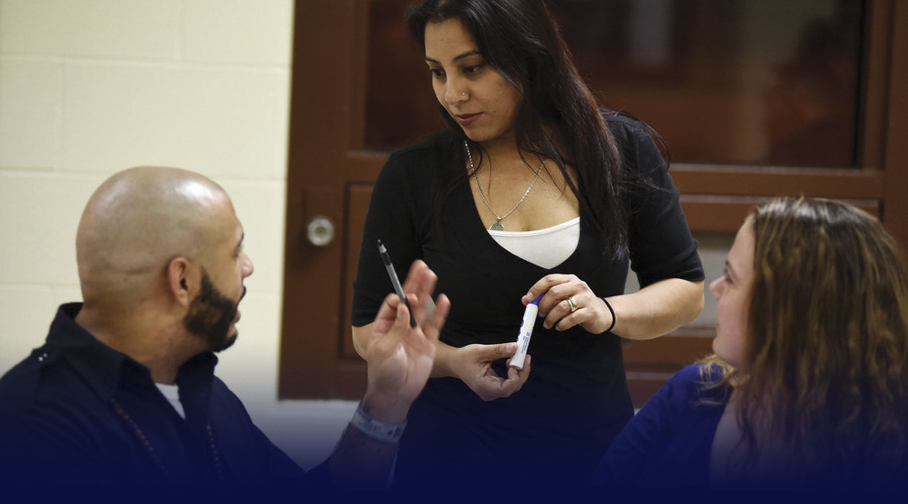 Assistant teaching professor, Shaheen Pasha, standing and holding a blue dry erase marker, in discussion with two students.