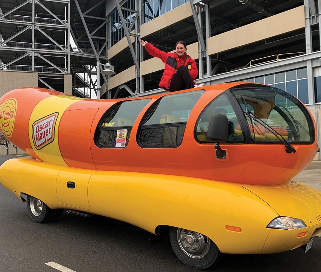 A student sits on top of the Oscar Mayer weinermobile - which is a car shaped like a hot dog.