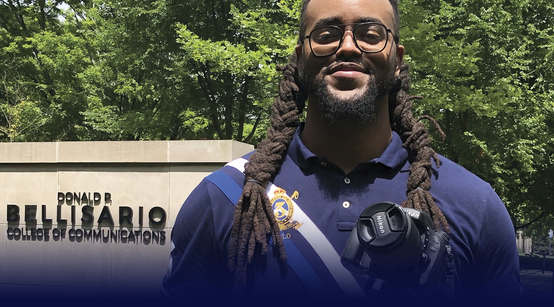 Alumnus Paris Palmer, now strategic communications coordinator for the
Bellisario College, poses with a camera in front of a wall on campus.