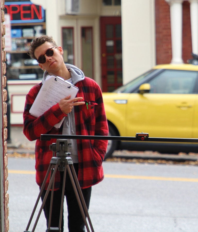 A student in sunglasses and a red and black plaid overcoat stands behind a camera on a city street