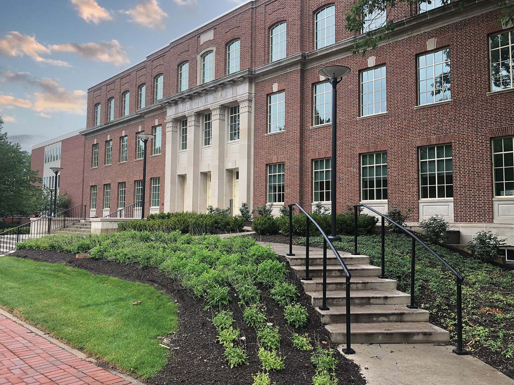 A subtle background photo of the front of Willard Building, a several story brick classroom building with four columns at the ntrance. Green lawn and bushes in the foreground.