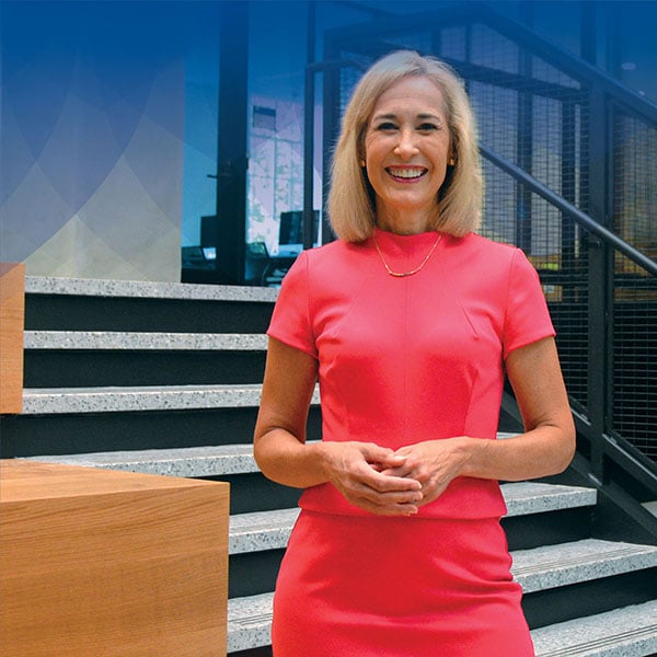 Marie Hardin, Dean of the Bellisario College is smiling with shoulder length blonde hair and is dressed in a bright coral dress. She is posing in the new Bellisario Media Center with a flight of concrete stairs behind her.