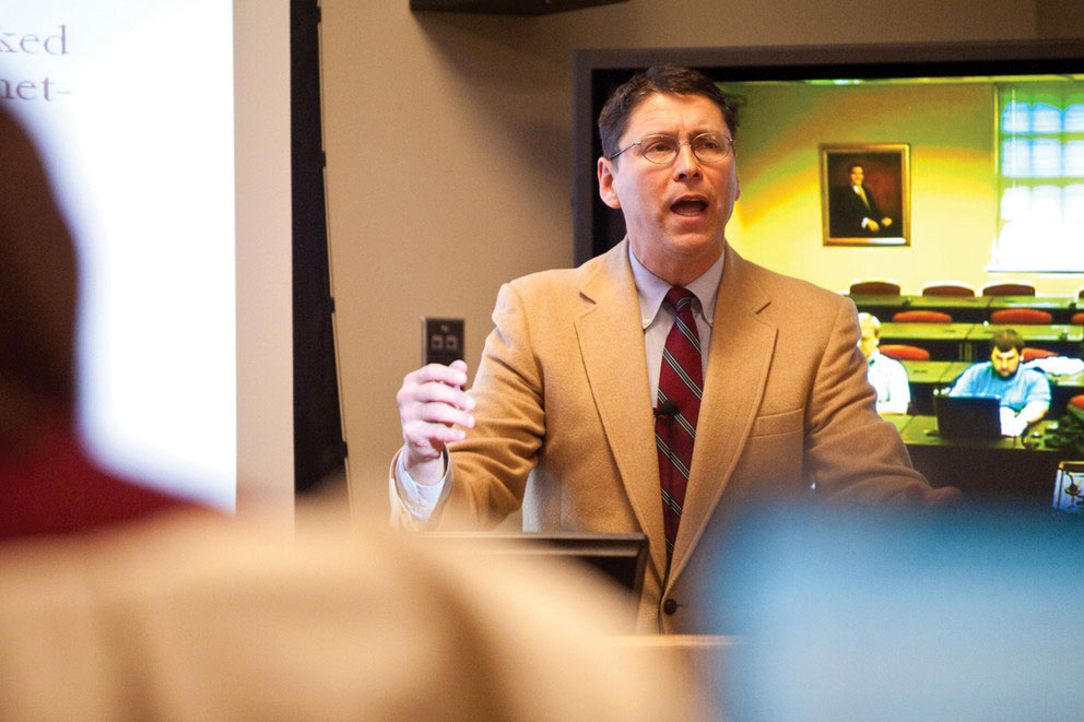 Professor Rob Freiden, wearing a tan suit jacket and glasses, lectures in front of a classroom.