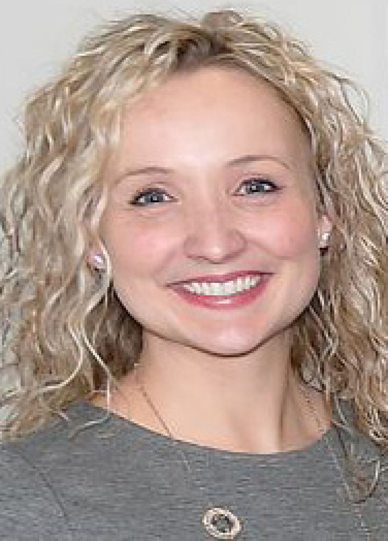 A woman with wavy blonde hair wearing a grey sweater smiles broadly.
