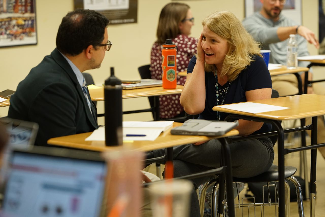 a man with dark hair and glasses wearing a suit talks with a woman with blonde hair and a navy blue top