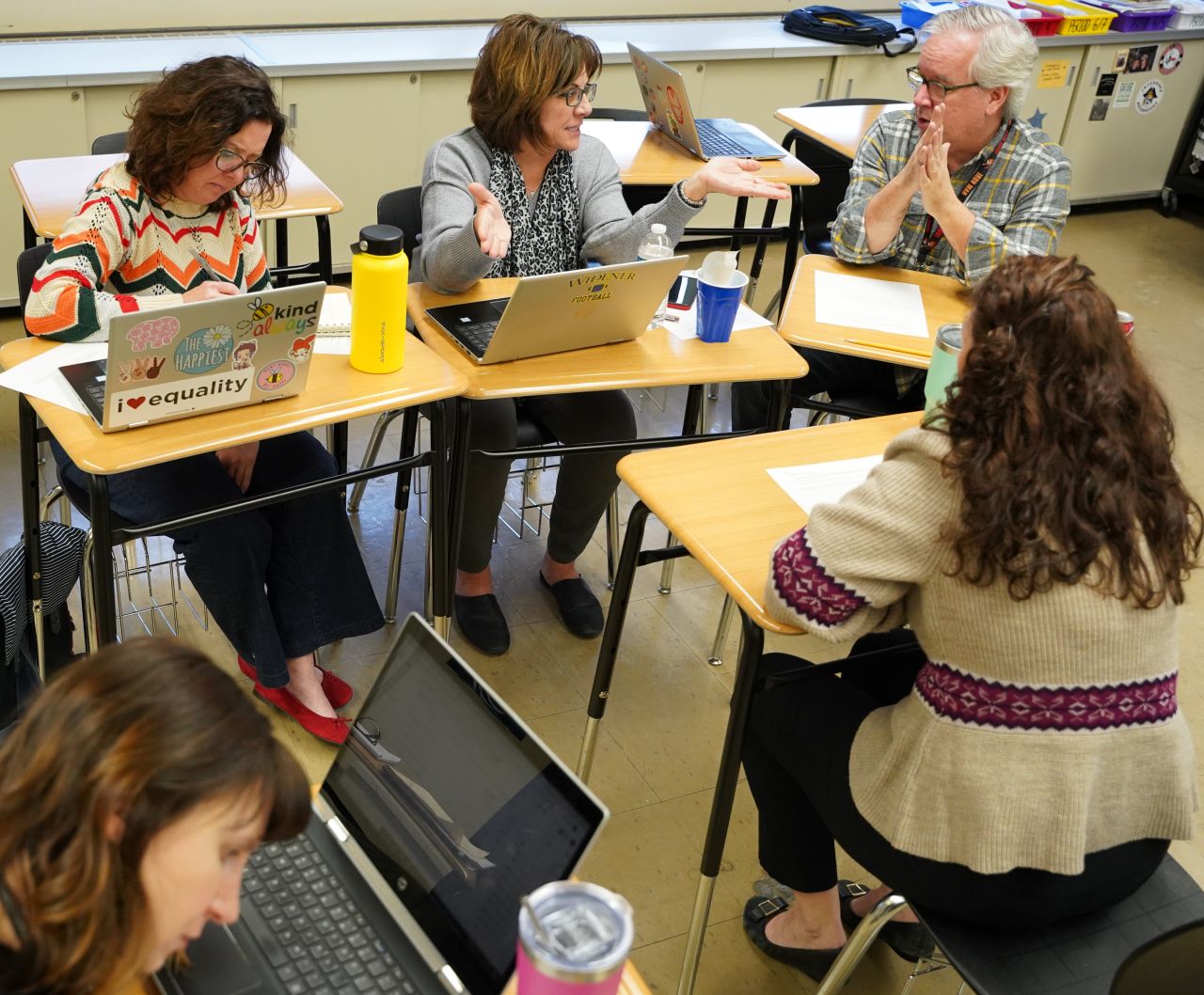 Four people sit at desks in a circle talking; three women all with short brown hair and one man with gray hair and glasses