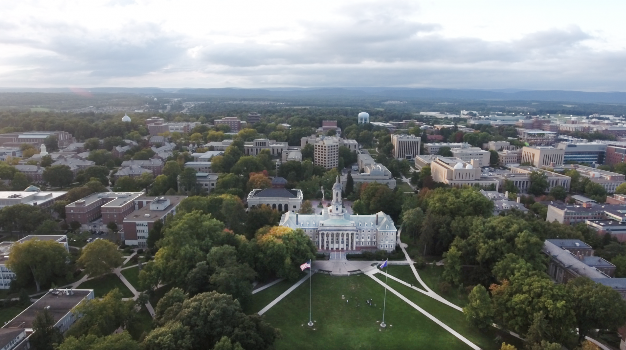 Aerial shot of Penn State University's main campus with the Old Main building (white stone) pictured in the center with two flag poles surrounded by tree-lined paths and other academic buildings.