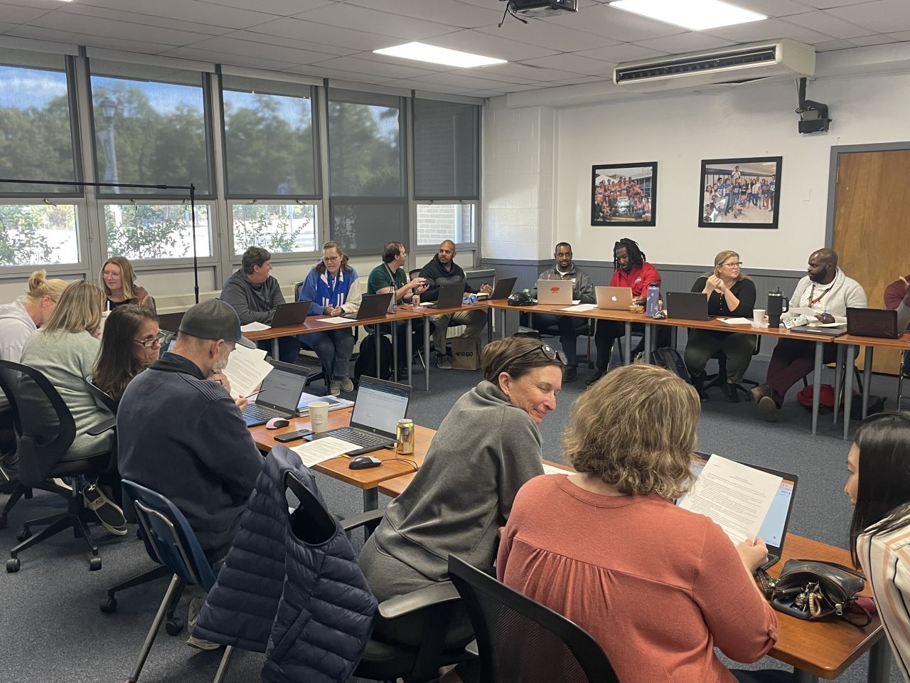 A room full of female, male, Caucasian and African-America people sit in a horseshoe shape with laptops discussing an article