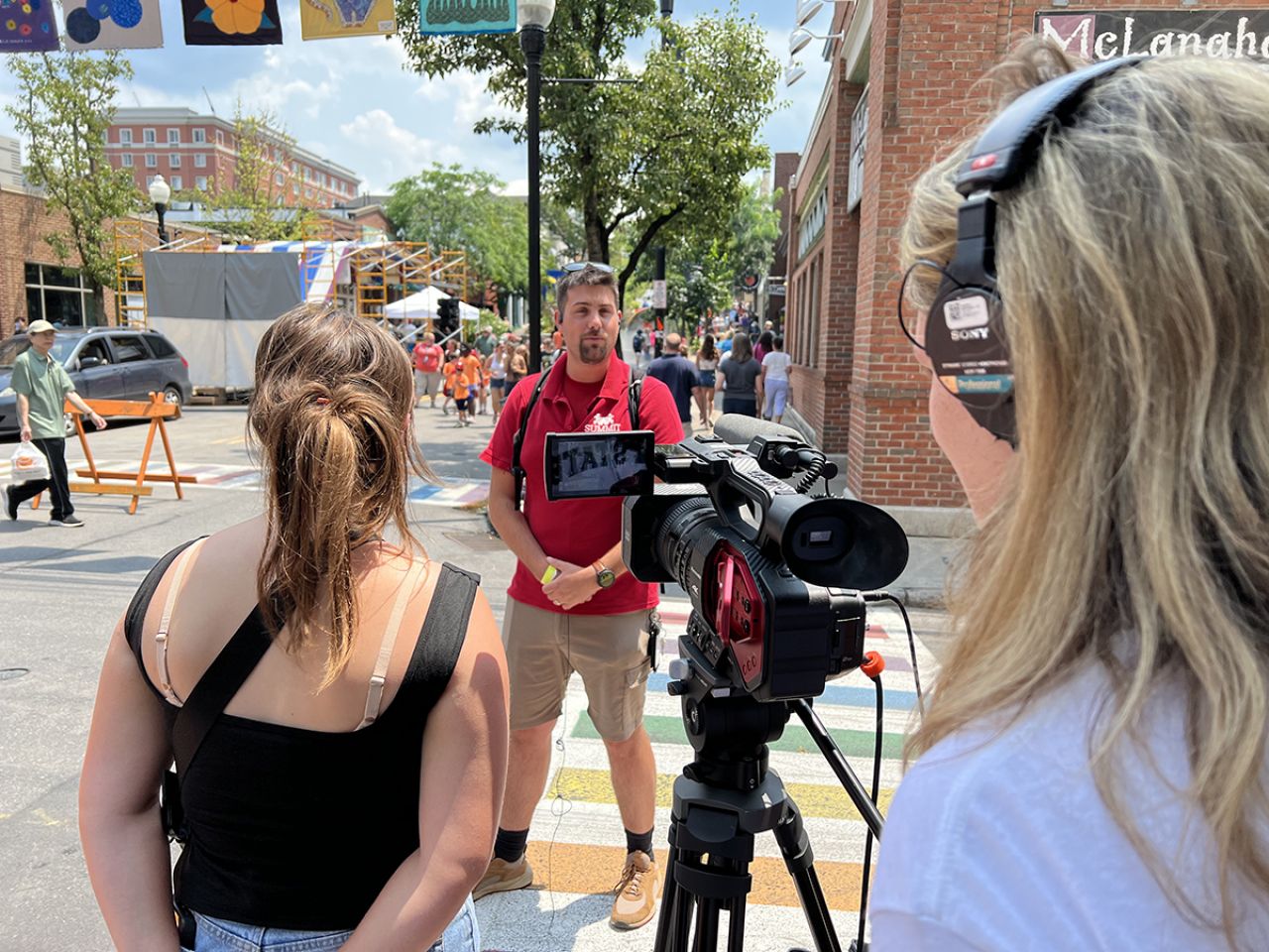 A gentleman in the center of the screen gets interviewed by two students with a camera who are positioned left and right on the image.