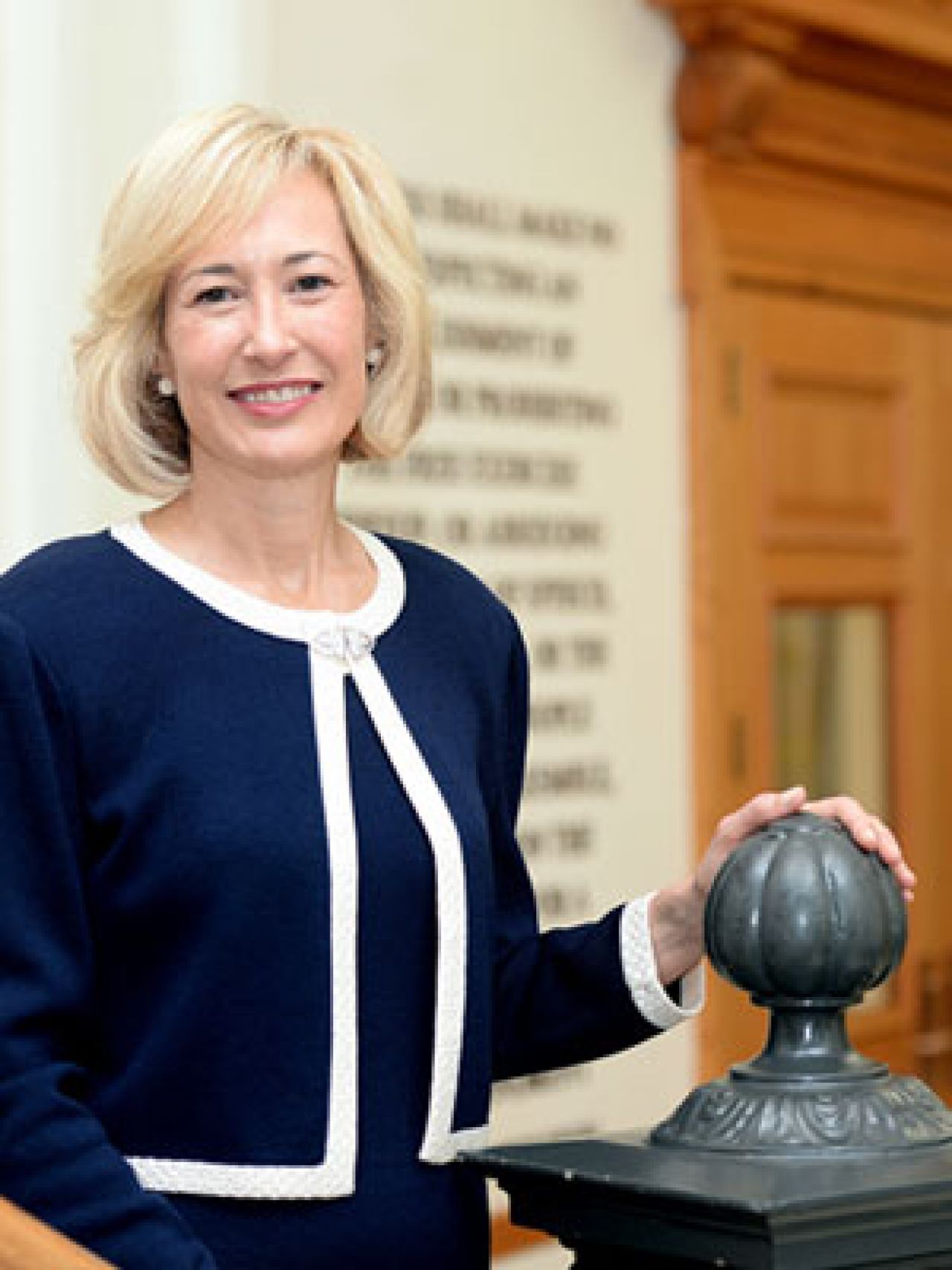 Dean Marie Hardin, a woman with shoulder-length blonde hair wearing a blue pant suit, stands in a building lobby with an impressive wooden doorway in the background.