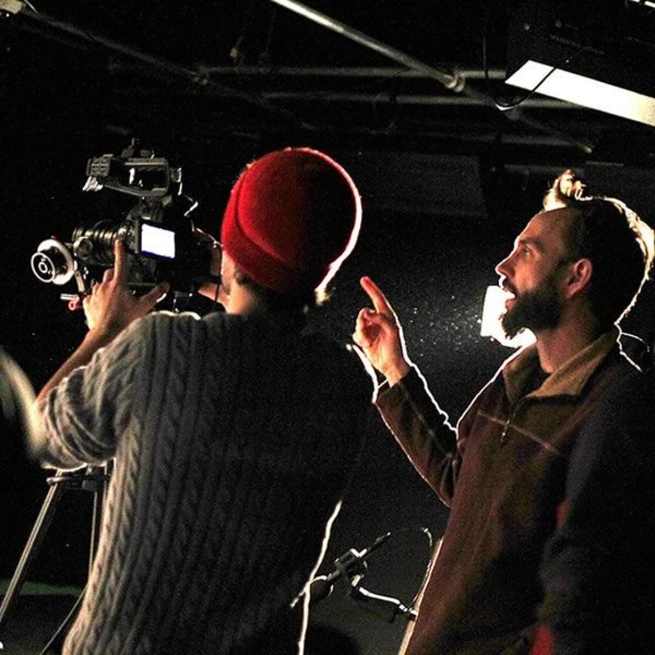 A student in cable knit sweater and red beanie cap holds a camera while receiving instruction from a bearded faculty member on the right.