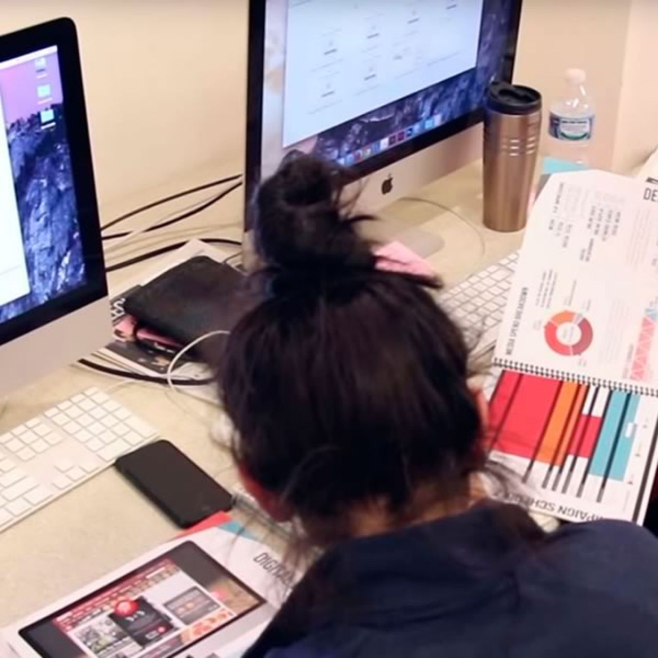 A female advertising student sits in front of a computer working on a presentation. We see her dark brown pulled up in a pony tail and some printed report pages in front of her with graphs and creative screenshots.