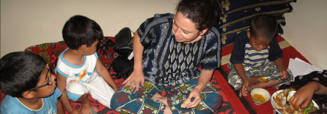 A woman dressed in a patterned black and white shirt sits among three children while they eat a meal.