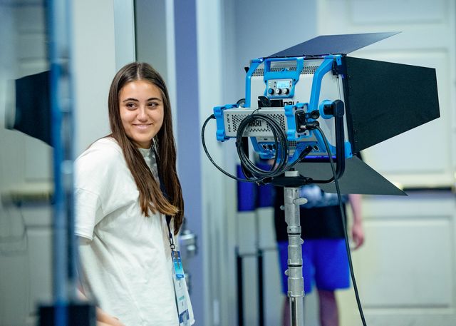 A student smiles at the camera while standing next to a light kit.