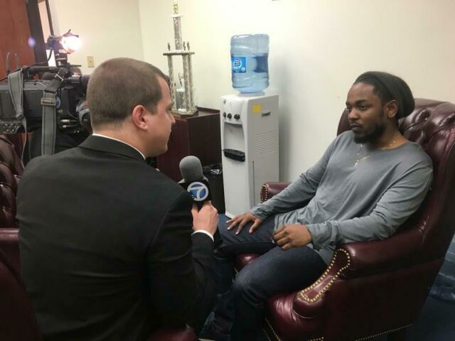 A student wearing a suit and holding a microphone interviews rapper Kendrick Lamar, sitting in a leather chair and wearing a gray shirt and slouchy beanie cap