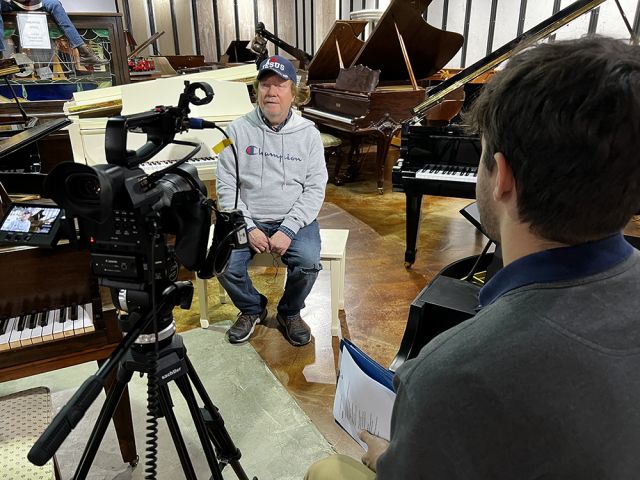Man sitting amongst pianos during an on-camera interview.