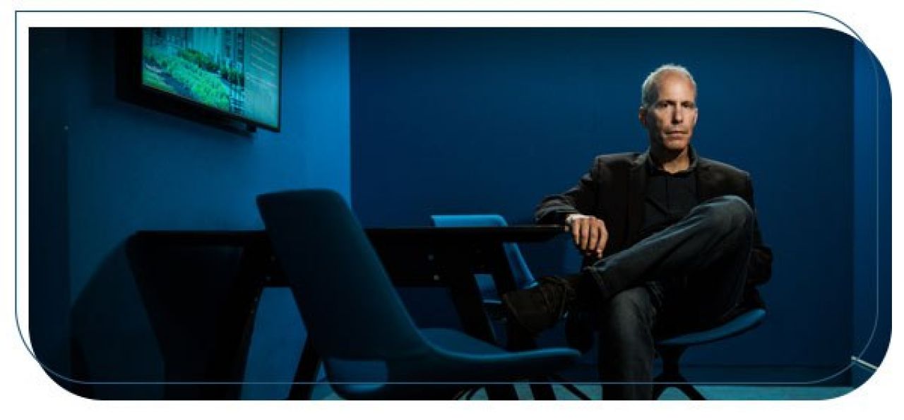 A man in dark pants and a black top and blazer is seated in a chair in a dark blue room