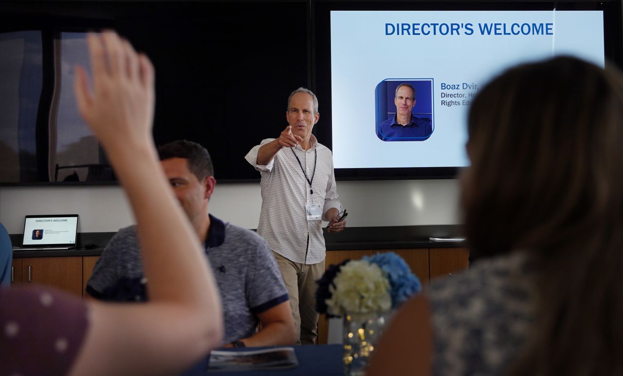 man in a white button up shirt points at the camera while speaking to a group