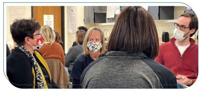 Three women and one man are seated in chairs in a circle in a classroom, wearing masks and having a conversation.