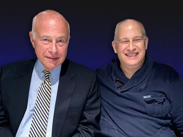 two older men sit next to each other; man on the left is wearing a dark suit, blue shirt, and yellow tie; man on the right is wearing a navy blue sweater