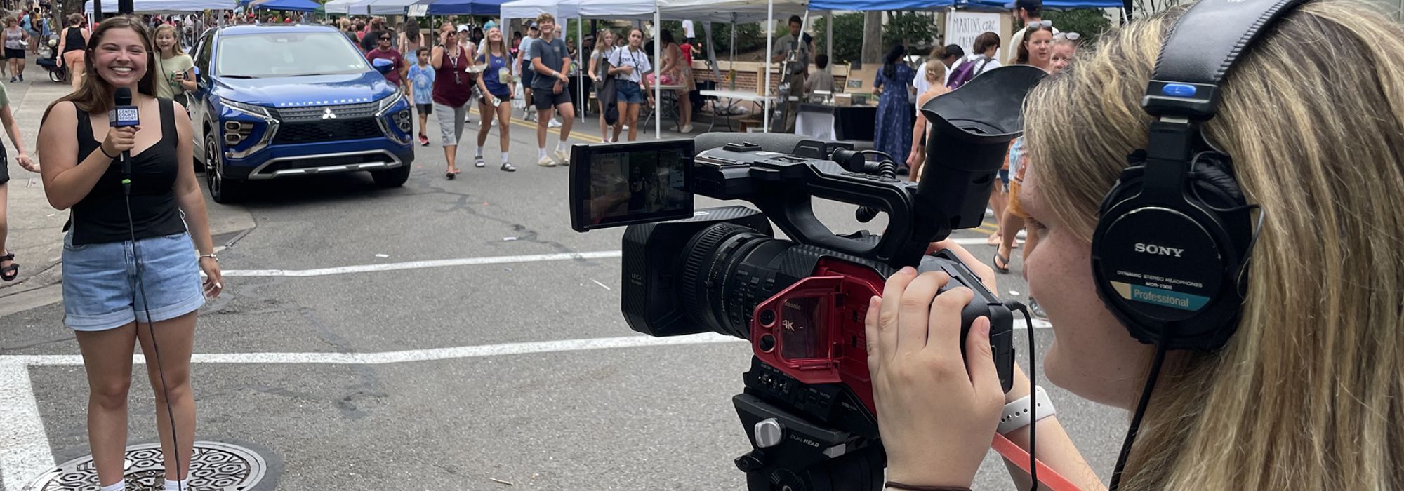 A student on the left side of the photo holds a microphone while a student on the right side directs the camera in her direciton.