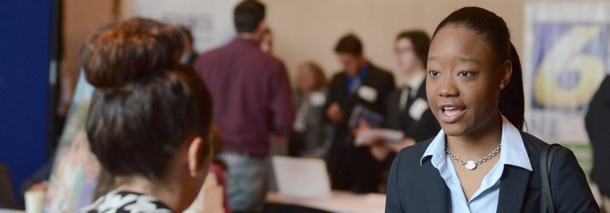 A student smartly dressed in a blue blazer and button-down talks with a recruiter at a job fair.