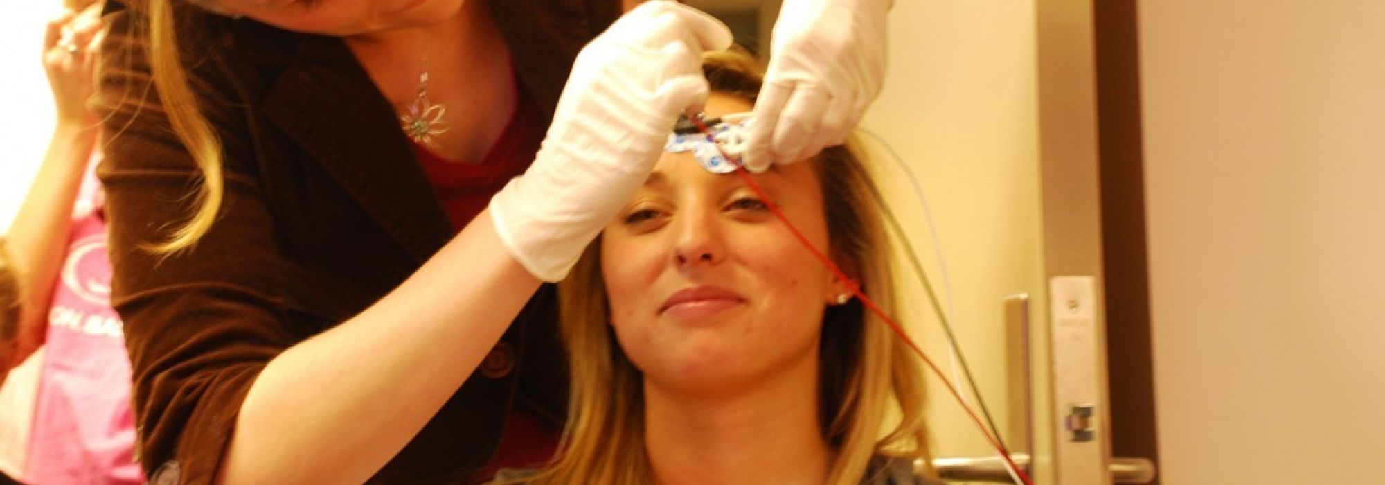 A woman wearing medical gloves places a sensor on the forehead of a student sitting in a chair during the filming of an engineering experiment.