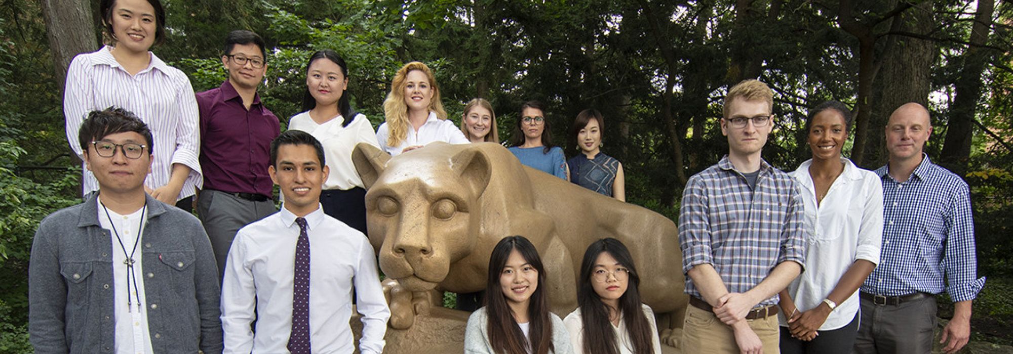 Fourteen graduate students pose in front of the Nittany Lion shrine, a large sculpture of a mountain lion carved out of limestone.
