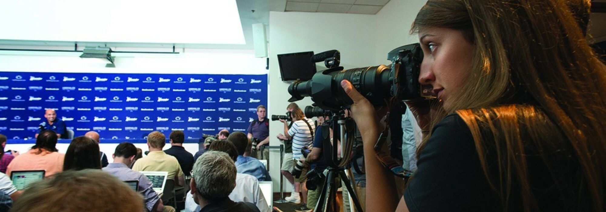 A student photographer for the Curley Center in the foreground is taking a photo of Penn State football coach James Franklin at a press conference.