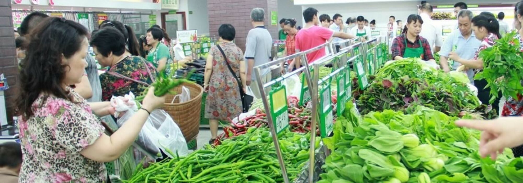 A bunch of shoppers around an open-air market look over fresh vegetables.