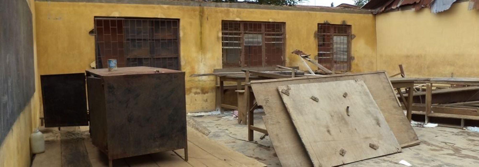 A classroom with yellow walls appears to have had its roof ripped off from a natural disaster. Scattered wood and desk remnants on the floor.