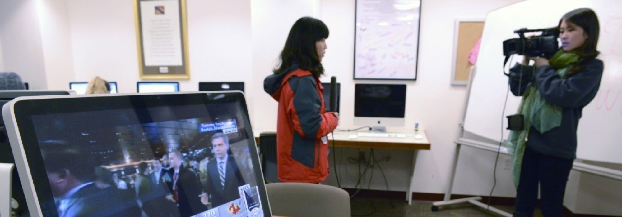 Two students in a conference room filming a news segment. On the left is a student wearing a red and blue rain coat and talking into a microphone. On the right is a student wearing a blue sweater and bright green scarf operating a video camera.