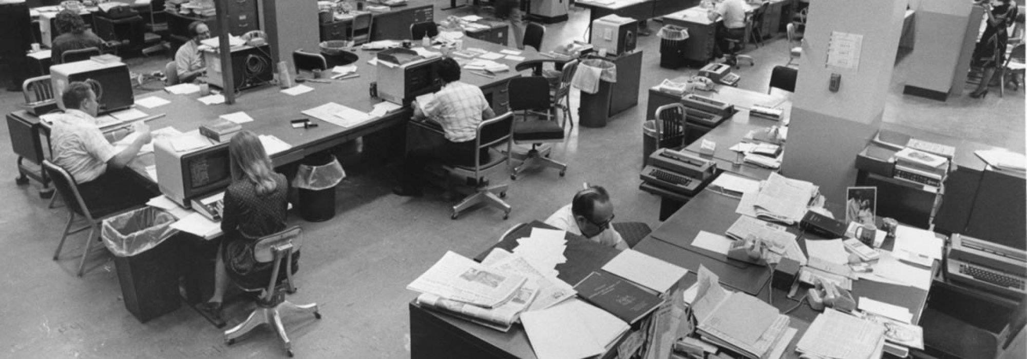 Black and white image shows an old newspaper newsroom with reporters surrounded by stacks of papers, sitting at old swiveling desk chairs, working at 1970s era computers.