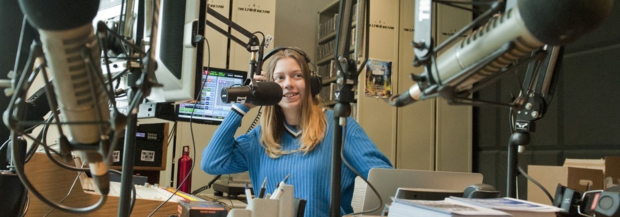 A blonde female student wearing a blue sweater stands surrounded by microphones in a recording studio