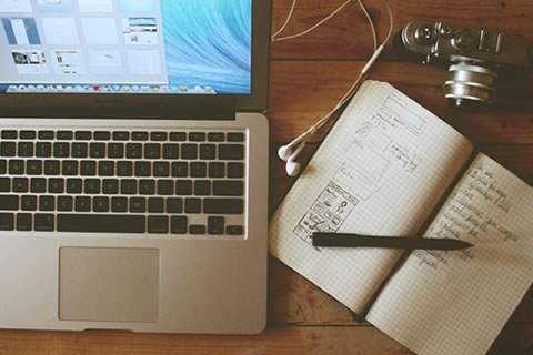 An open laptop and notebook along with a camera sit on a desk