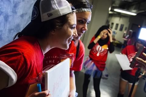 Two students wearing baseball caps backwards hold an encouraging sign for THON participants towards a camera person