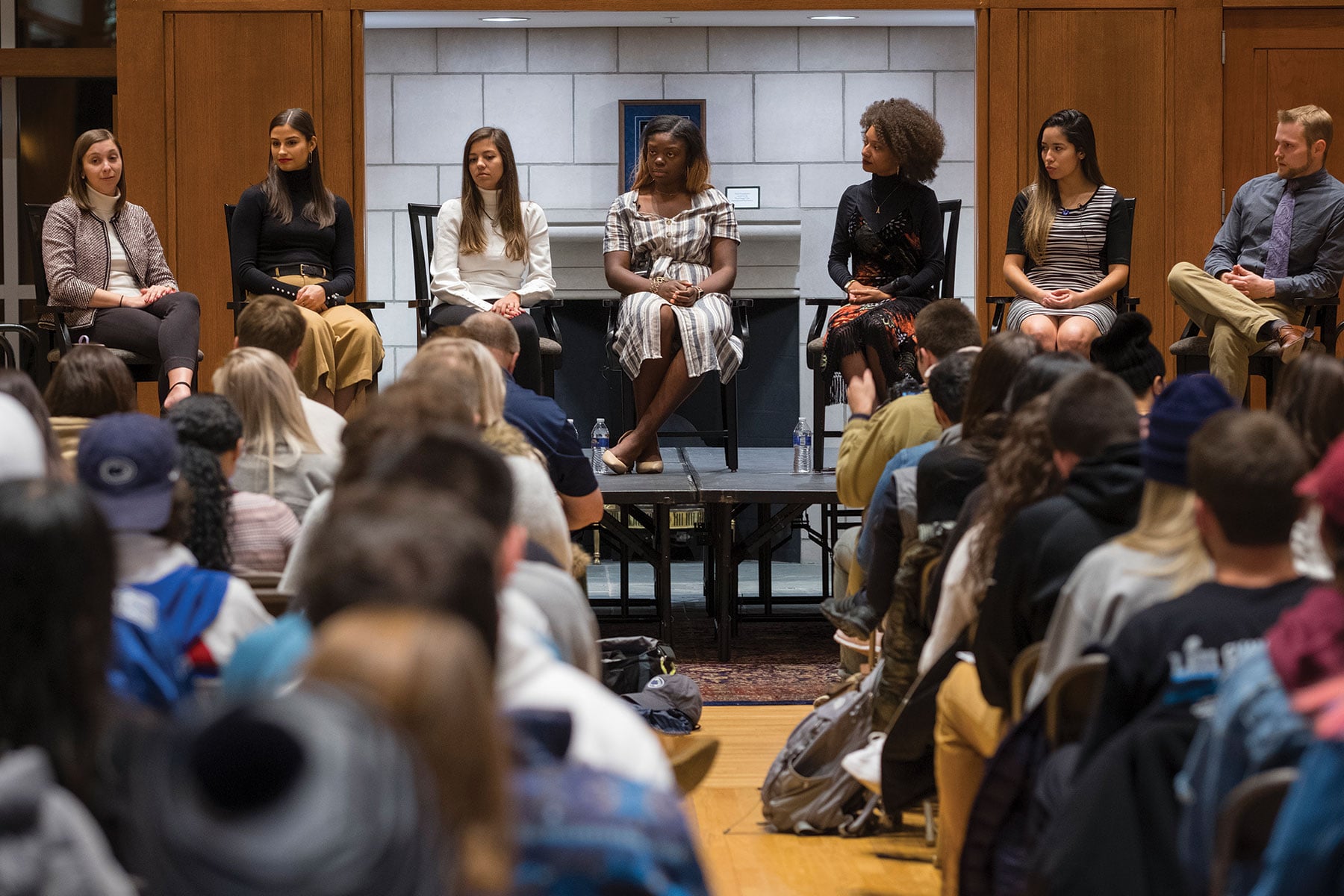 A panel of seven young alumni sit on a stage in front of a student group answering questions