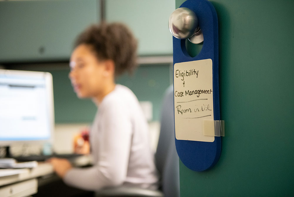 Super soft focus background of a woman on a laptop. In the foreground in focus is a door hangar that reads - Eligibility and Case Management - Room in Use