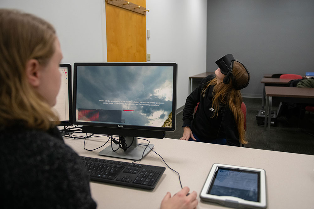 A student wearing VR goggles looks up towards the ceiling while another student gives directions from behind her computer.