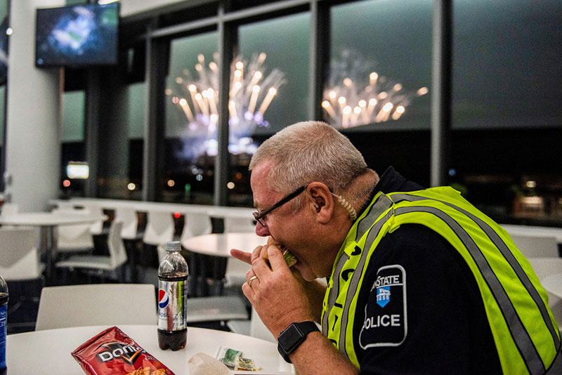 A police officer eats a sandwich as post-game fireworks light up the sky outside.