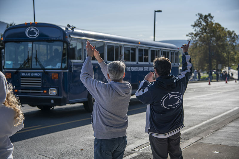 Fans clapping and waving at a blue school bus that transports the Penn State football team to the stadium.