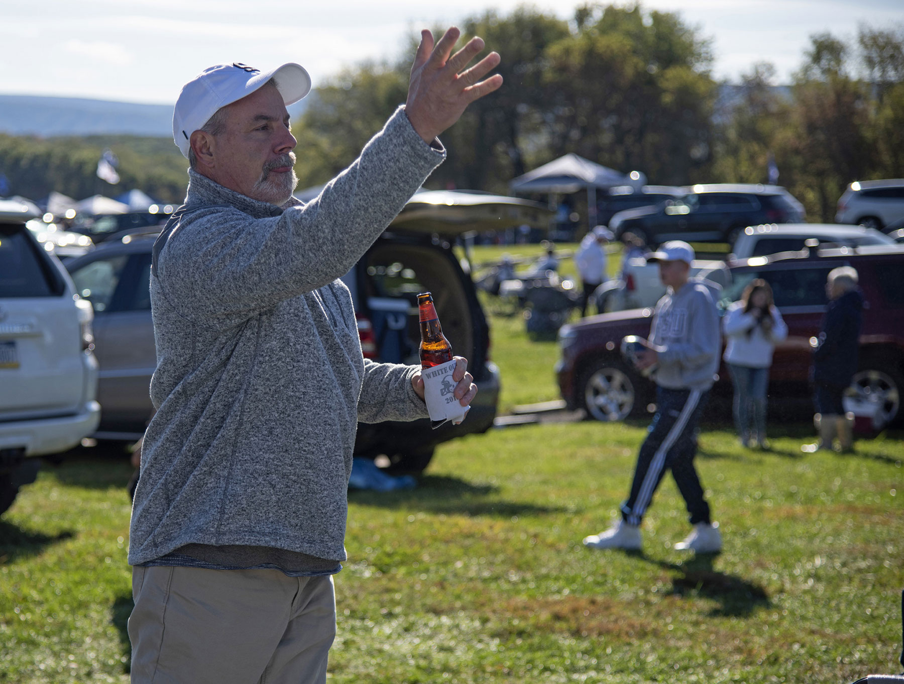 A fan in gray sweatshirt and white Penn State baseball cap tosses a cornhole bag.
