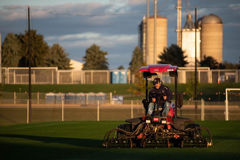 A groundskeeper mows a soccer field with farming silos in the background.