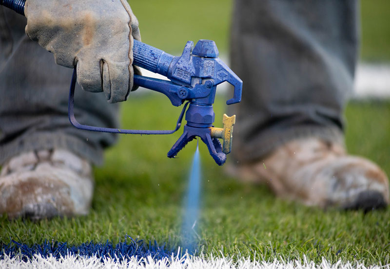 Closeup of a paint sprayer putting down blue paint on the grass at Beaver Stadium.