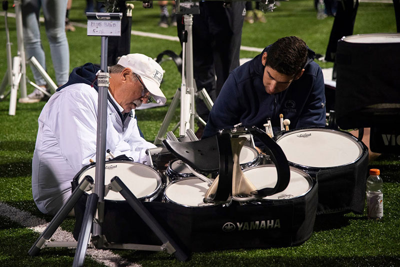 Dave Cree crouches with a drummer as they examine a broken drum.