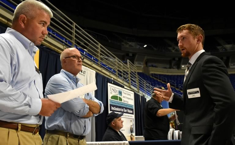 Zack dressed ina= a suit talking to recruiters at a career fair.